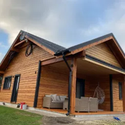 Nowoczesny, parterowy dom z drewna o jasnej elewacji, z zadaszonym tarasem wyposażonym w rattanowe meble wypoczynkowe i wiszący fotel, widok z zewnątrz na tle pochmurnego nieba.