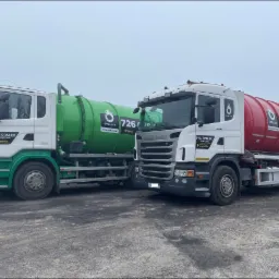 Dwa samochody ciężarowe Scania z cysternami: jeden zielony, drugi czerwony, oba z logo firmy PHU Opałek i numerem telefonu, stojące na szarym, betonowym placu w pochmurny dzień.