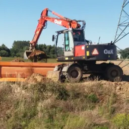 Pomarańczowa koparka O&K ładuje ziemię do pomarańczowej wywrotki na tle pola i słupa wysokiego napięcia.