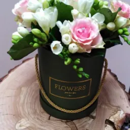 Kompozycja kwiatowa w okrągłym, ciemnozielonym pudełku z napisem 'FLOWERS' w złotej ramce, umieszczona na drewnianym stoliku z widocznymi słojami.