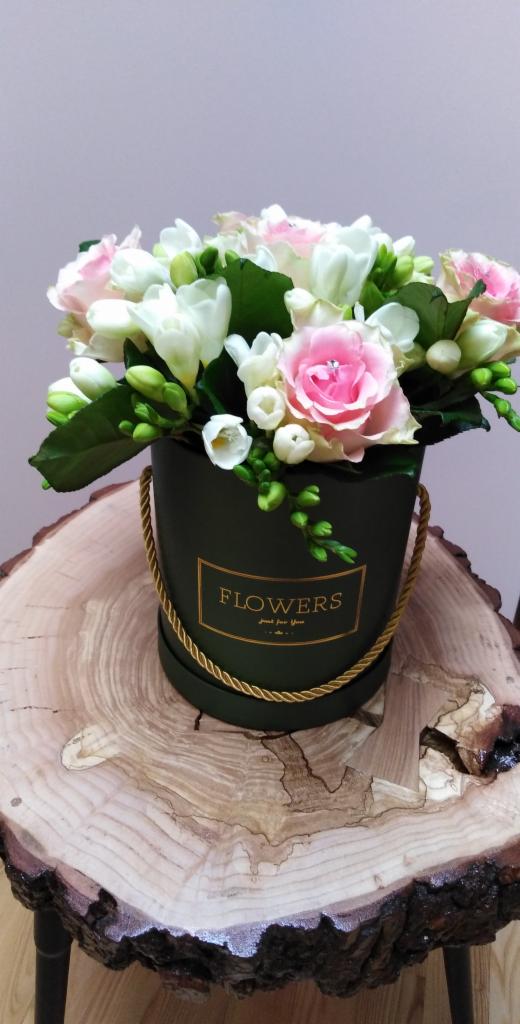 Kompozycja kwiatowa w okrągłym, ciemnozielonym pudełku z napisem 'FLOWERS' w złotej ramce, umieszczona na drewnianym stoliku z widocznymi słojami.