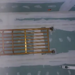 Złoty, metalowy grzejnik łazienkowy zamontowany na ścianie z płyt kartonowo-gipsowych, widoczny poziomica. Ściana przygotowana do dalszych prac wykończeniowych.