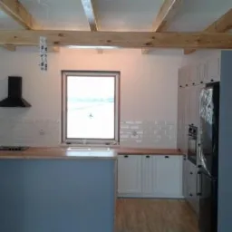 Wnętrze kuchni w trakcie montażu: widoczne białe szafki z drewnianymi blatami, wyspa kuchenna w kolorze szarym, czarny okap, drewniane belki stropowe, drabina oparta o ścianę.