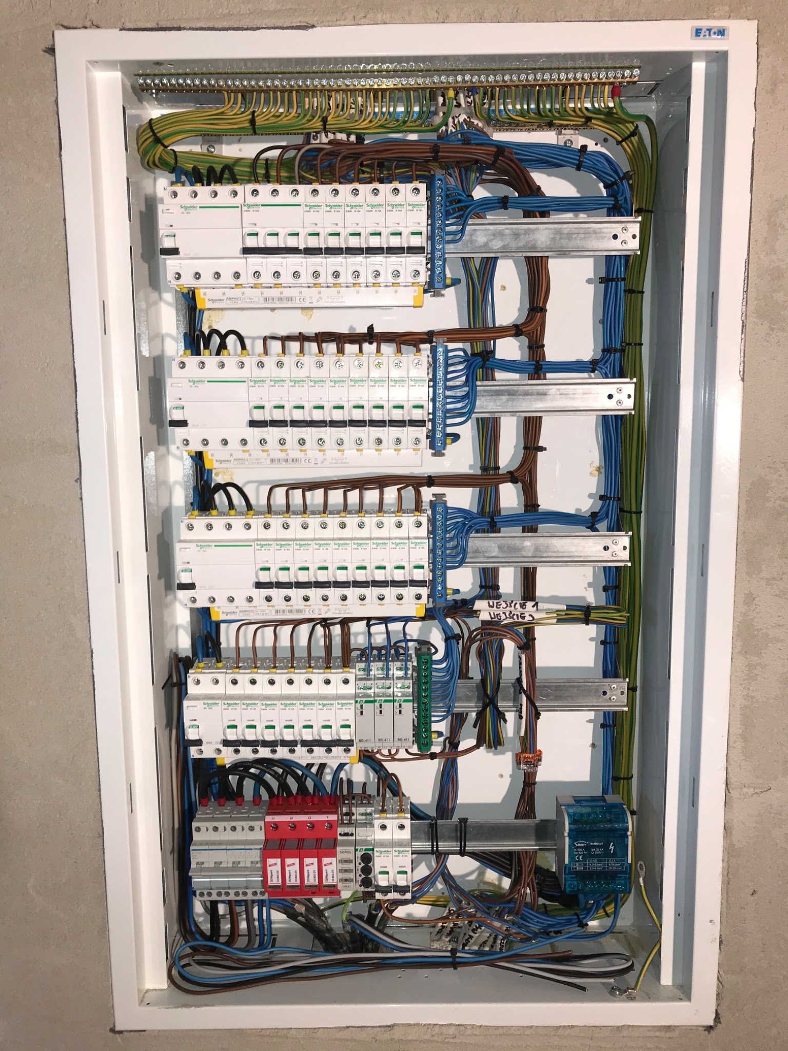 Rozdzielnica elektryczna z zainstalowanymi bezpiecznikami, zabezpieczeniami przepięciowymi i uporządkowanymi przewodami w kolorach: żółto-zielonym, niebieskim i brązowym, zamontowana w białej...