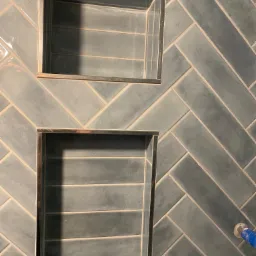 Niebiesko-szare, błyszczące płytki ułożone w jodełkę na ścianie z wnękami wykończonymi metalową listwą.