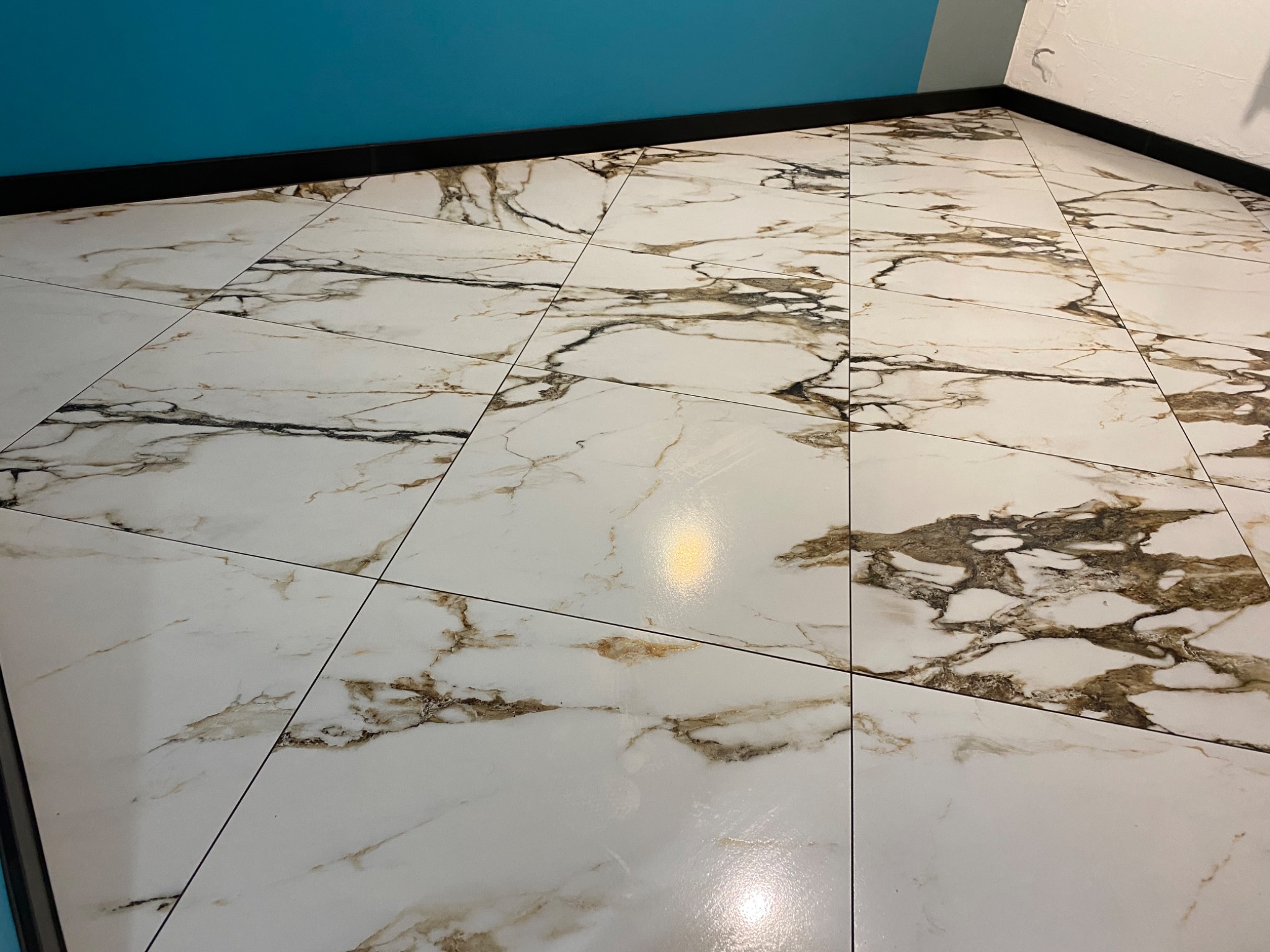 Błyszcząca podłoga z białych płytek imitujących marmur z brązowymi żyłami, ułożona w kwadraty w pomieszczeniu w Zielonej Górze z turkusową ścianą u góry.