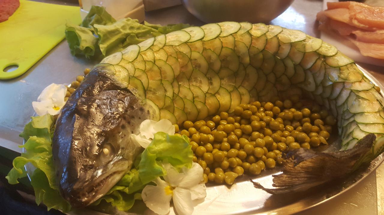 Ryba w galarecie, udekorowana plastrami ogórka, groszkiem konserwowym, sałatą i białymi kwiatami, podana na srebrnej tacy.
