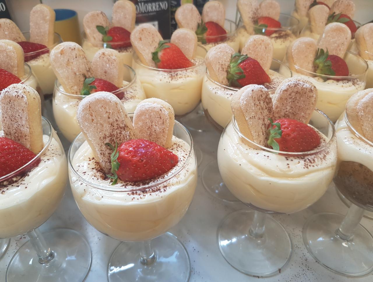 Deser w szklance na nóżce: kremowy pudding z truskawką i biszkoptami oprószonymi kakao, ustawiony w rzędach z innymi porcjami.