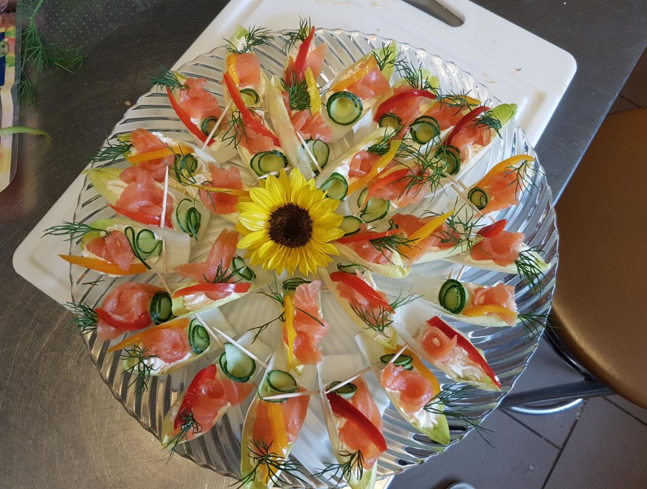 Dekoracyjna szklana patera z przekąskami: łódeczki z cykorii z kremowym serem, łososiem, ogórkiem i papryką, ozdobiona słonecznikiem w centrum.