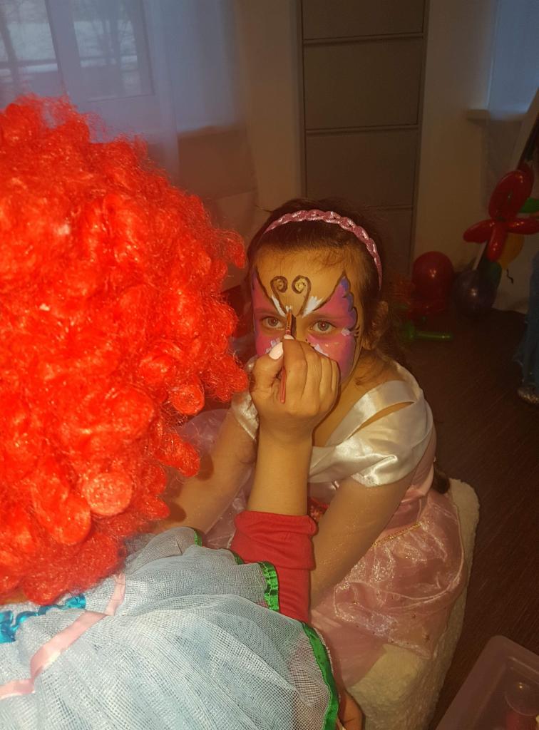 Dziewczynka w różowej sukience z motylkowym makijażem na twarzy, malowana przez osobę w czerwonej peruce.