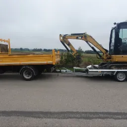 Żółty samochód ciężarowy Iveco z zabudową skrzyniową ciągnie przyczepę z mini koparką CAT 301.5L, na tle pola i pochmurnego nieba.