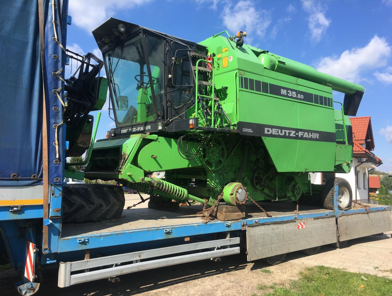 Zielony kombajn Deutz-Fahr M35.80 zabezpieczony łańcuchami na niebieskiej platformie transportowej pod błękitnym niebem.