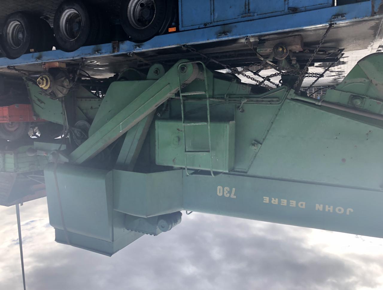 Transport niskopodwoziowy: Zielona maszyna rolnicza John Deere 730 zabezpieczona łańcuchami na niebieskiej platformie transportowej, widoczne koła platformy.