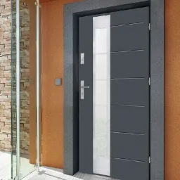 Szare drzwi wejściowe z pionowym przeszkleniem, osadzone w ciemnej, kamiennej futrynie, z widocznym zamkiem i klamką, obok szklanej ściany i beżowej elewacji.