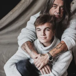 Portret ojca obejmującego syna w swetrach na tle płótna, widoczne tatuaże na ramionach syna i zegarek na jego nadgarstku.