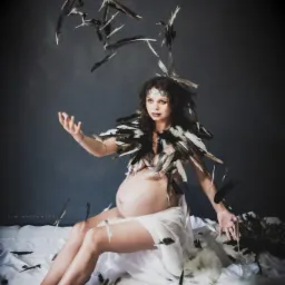 Kobieta w ciąży siedzi na białym materiale, ubrana w pióra, z piórami unoszącymi się w powietrzu, na ciemnoszarym tle.