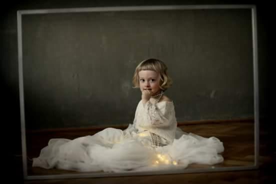 Dziewczynka w białej sukience i perłach pozuje w ramie na tle szarej ściany, oświetlona ciepłym światłem.
