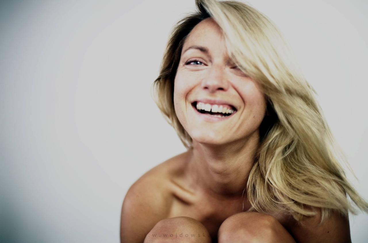 Portret uśmiechniętej kobiety z rozwianymi blond włosami na jasnym tle, uchwycony w naturalnym świetle.