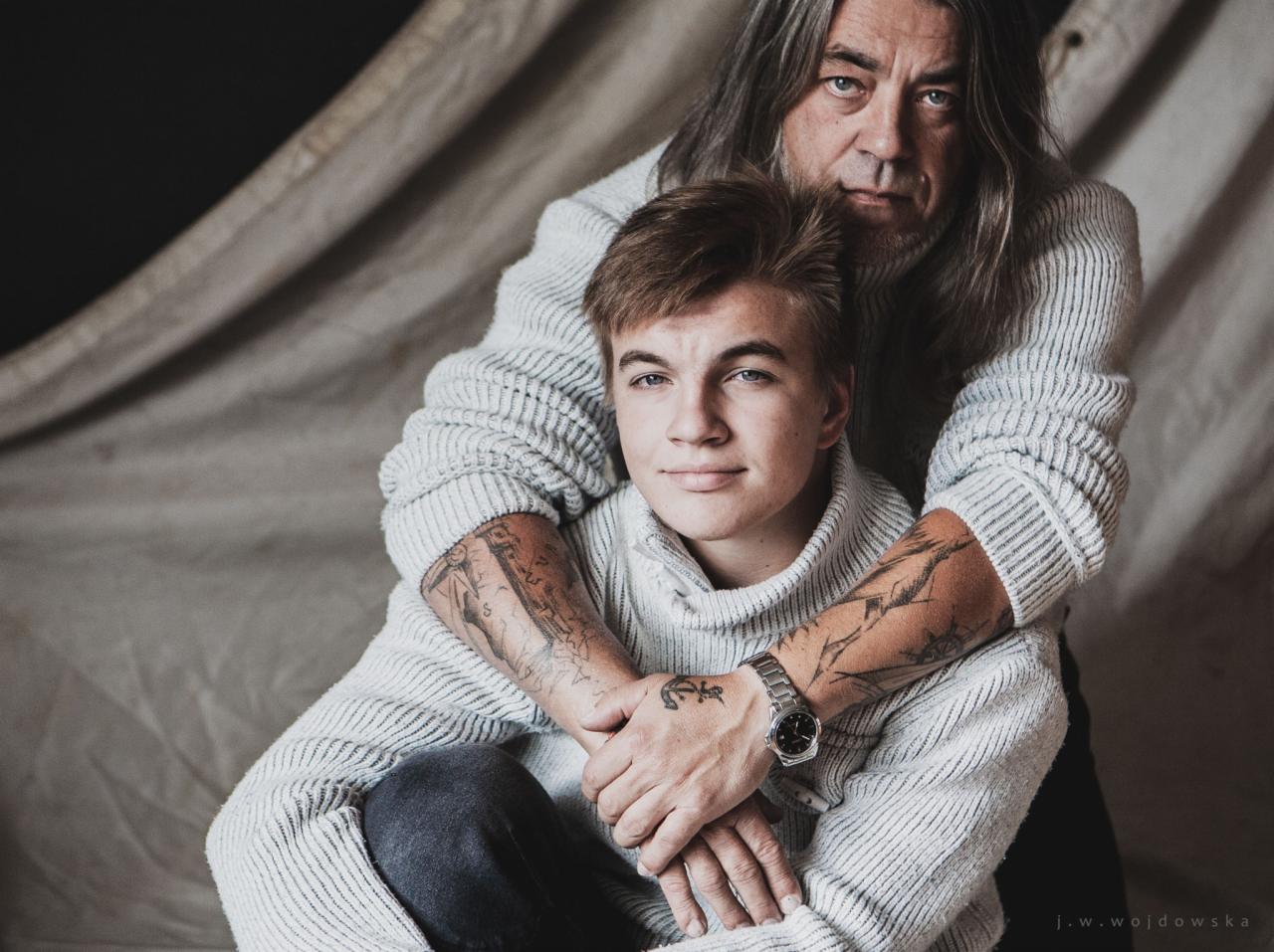 Portret ojca obejmującego syna w swetrach na tle płótna, widoczne tatuaże na ramionach syna i zegarek na jego nadgarstku.