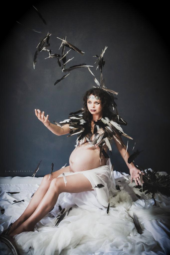 Kobieta w ciąży siedzi na białym materiale, ubrana w pióra, z piórami unoszącymi się w powietrzu, na ciemnoszarym tle.