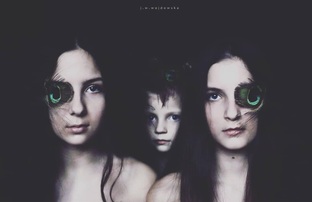 Portret artystyczny: dwie kobiety z piórami pawia zakrywającymi jedno oko, w tle dziecko z piórem we włosach, ciemne tło.