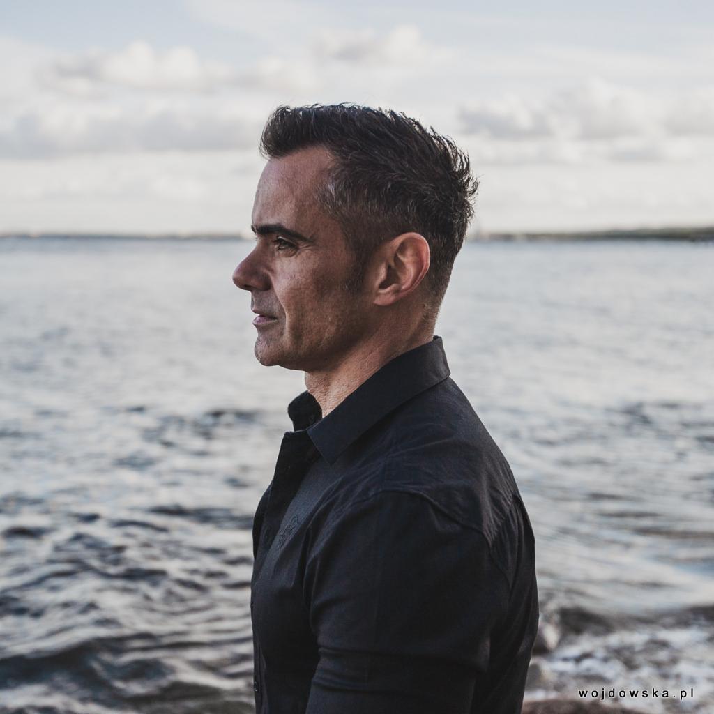 Portret biznesowy mężczyzny w czarnej koszuli na tle morza, profil, poważny wyraz twarzy, naturalne światło.
