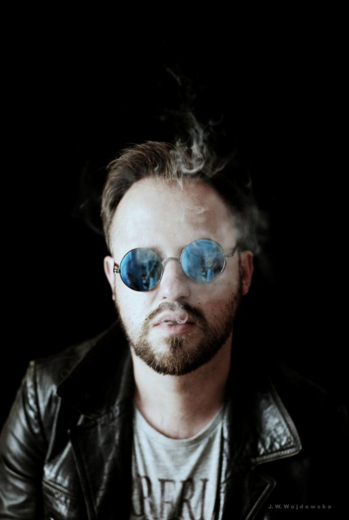 Portret mężczyzny z brodą w okrągłych okularach, z dymem unoszącym się z ust, ubrany w skórzaną kurtkę na tle czerni.