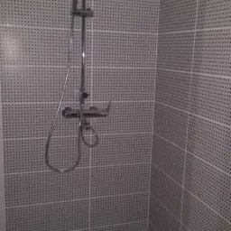 Kabina prysznicowa z szarymi płytkami w drobne czarne kropki i chromowaną armaturą, widoczny odpływ liniowy w podłodze.