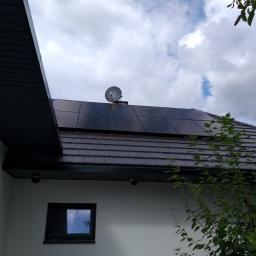 EKO-CARS BIS Bochenek Artur - Panele słoneczne na ciemnym dachu domu jednorodzinnego, widoczna antena satelitarna, fragment budynku z małym oknem i zielenią w tle.