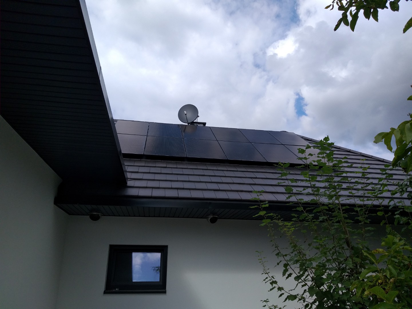Panele słoneczne na ciemnym dachu domu jednorodzinnego, widoczna antena satelitarna, fragment budynku z małym oknem i zielenią w tle.