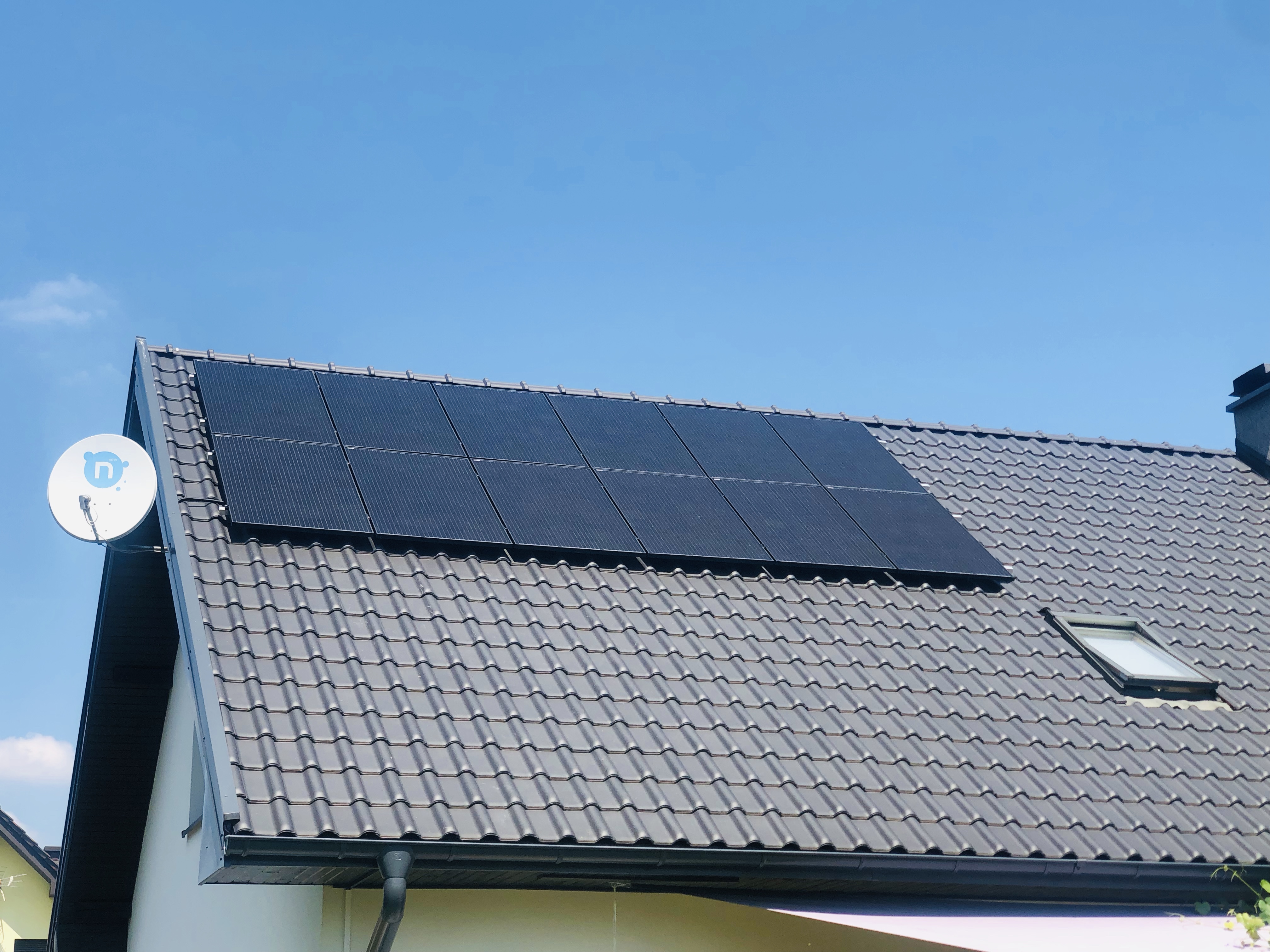 Panele słoneczne na szarym dachu z dachówką ceramiczną, widoczna antena satelitarna i okno dachowe, błękitne niebo w tle.