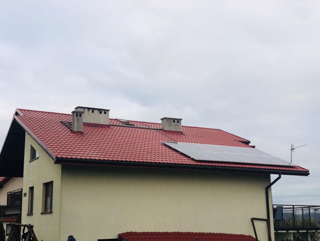 Panele słoneczne zamontowane na czerwonym dachu dwuspadowym budynku mieszkalnego z trzema kominami, widoczne z dołu w pochmurny dzień.