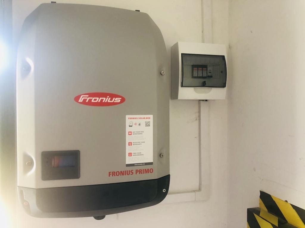 Szary falownik Fronius Primo zamontowany na białej ścianie obok skrzynki bezpiecznikowej, z widocznymi przewodami instalacji fotowoltaicznej.