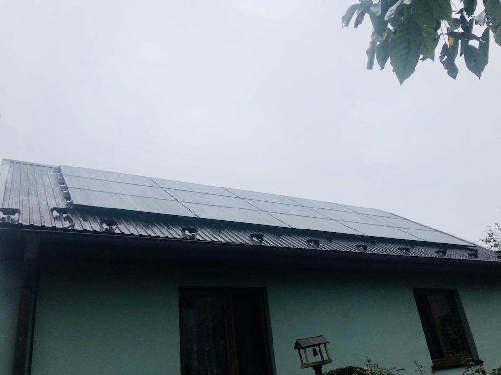 Panele słoneczne na metalowym dachu domu jednorodzinnego w pochmurny dzień, widoczne z poziomu trawnika, z ptasim domkiem w kadrze.