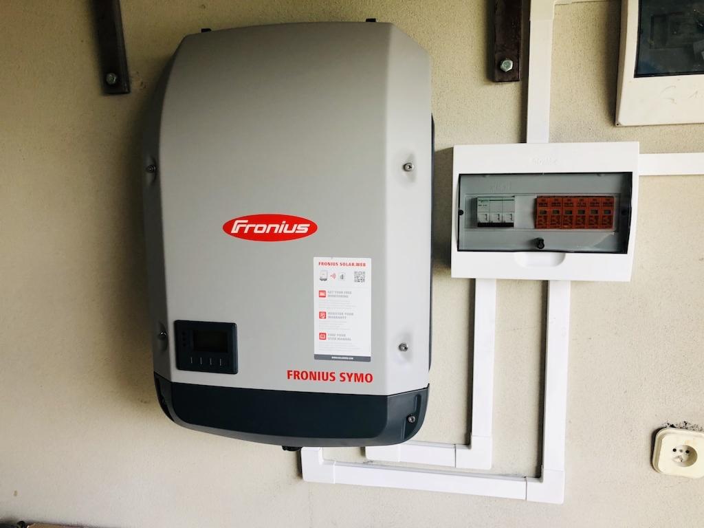 Szary falownik Fronius Symo zamontowany na ścianie obok skrzynki z bezpiecznikami i instalacji elektrycznej w białych rurkach.