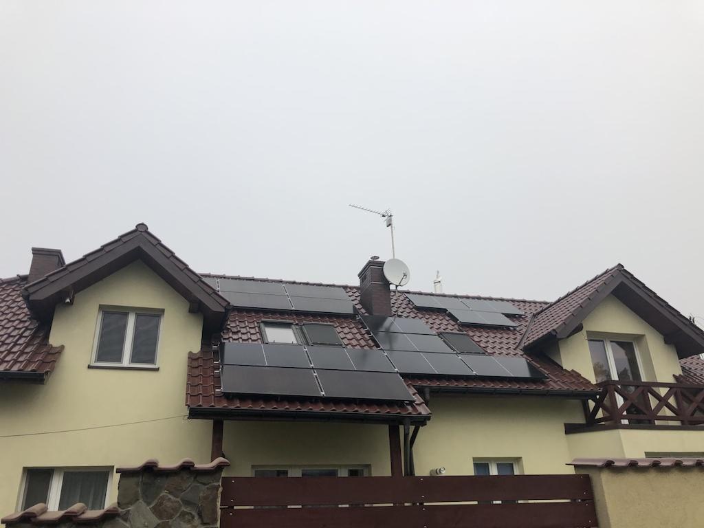 Panele słoneczne na dachu dwupiętrowego domu z brązową dachówką, widoczna antena satelitarna i komin.