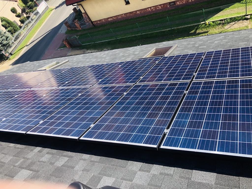 Panele słoneczne na dachu pokrytym szarym gontem bitumicznym, widok z góry na instalację i fragment posesji z budynkiem w tle.