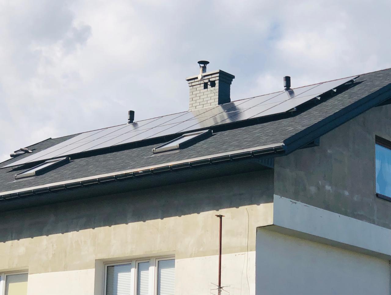 Panele słoneczne na dachu domu jednorodzinnego z szarym dachem i kominem z cegły, widoczne okna dachowe i fragment elewacji budynku.
