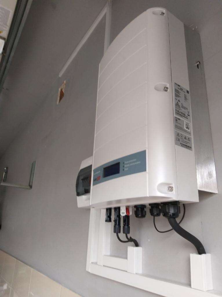 Biały falownik fotowoltaiczny zamontowany na szarej ścianie, widoczne kable połączeniowe i fragment instalacji elektrycznej.