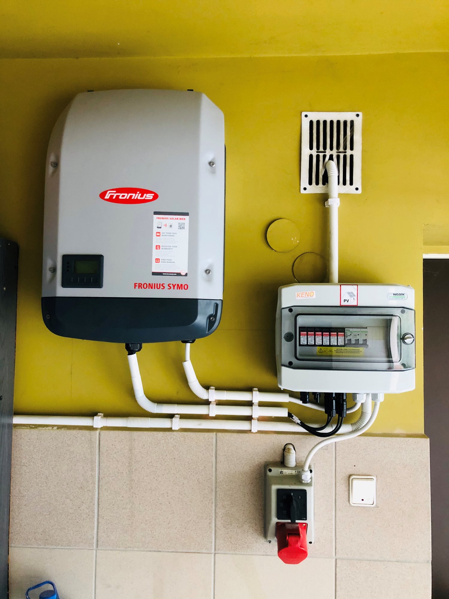 Szary falownik Fronius Symo z podłączonymi przewodami, obok skrzynka bezpiecznikowa Keno PV i wyłącznik prądu z czerwoną wtyczką na żółtej ścianie z kafelkami.