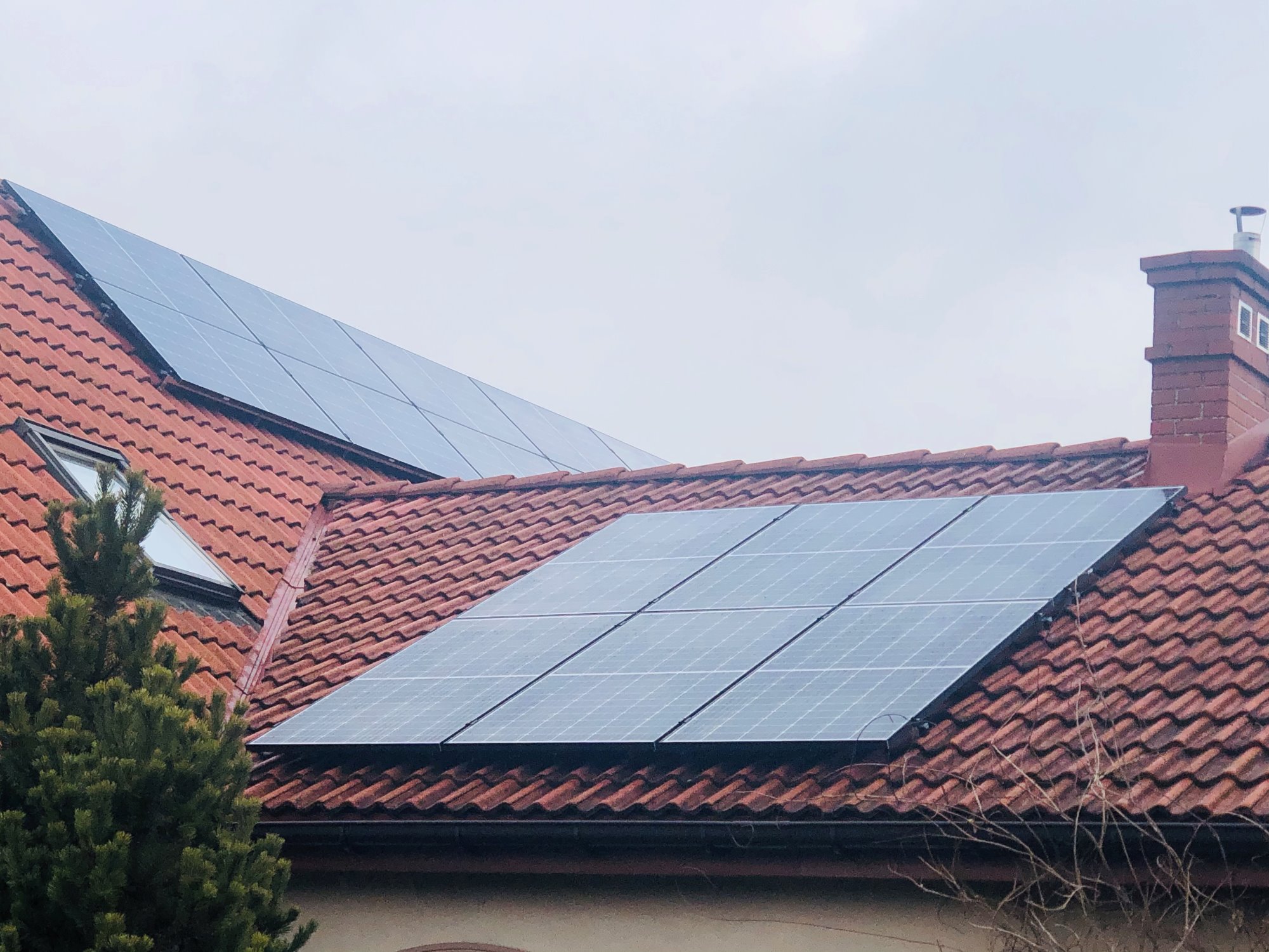 Panele słoneczne na dachu z czerwonej dachówki, widoczny komin i fragment drzewa iglastego.