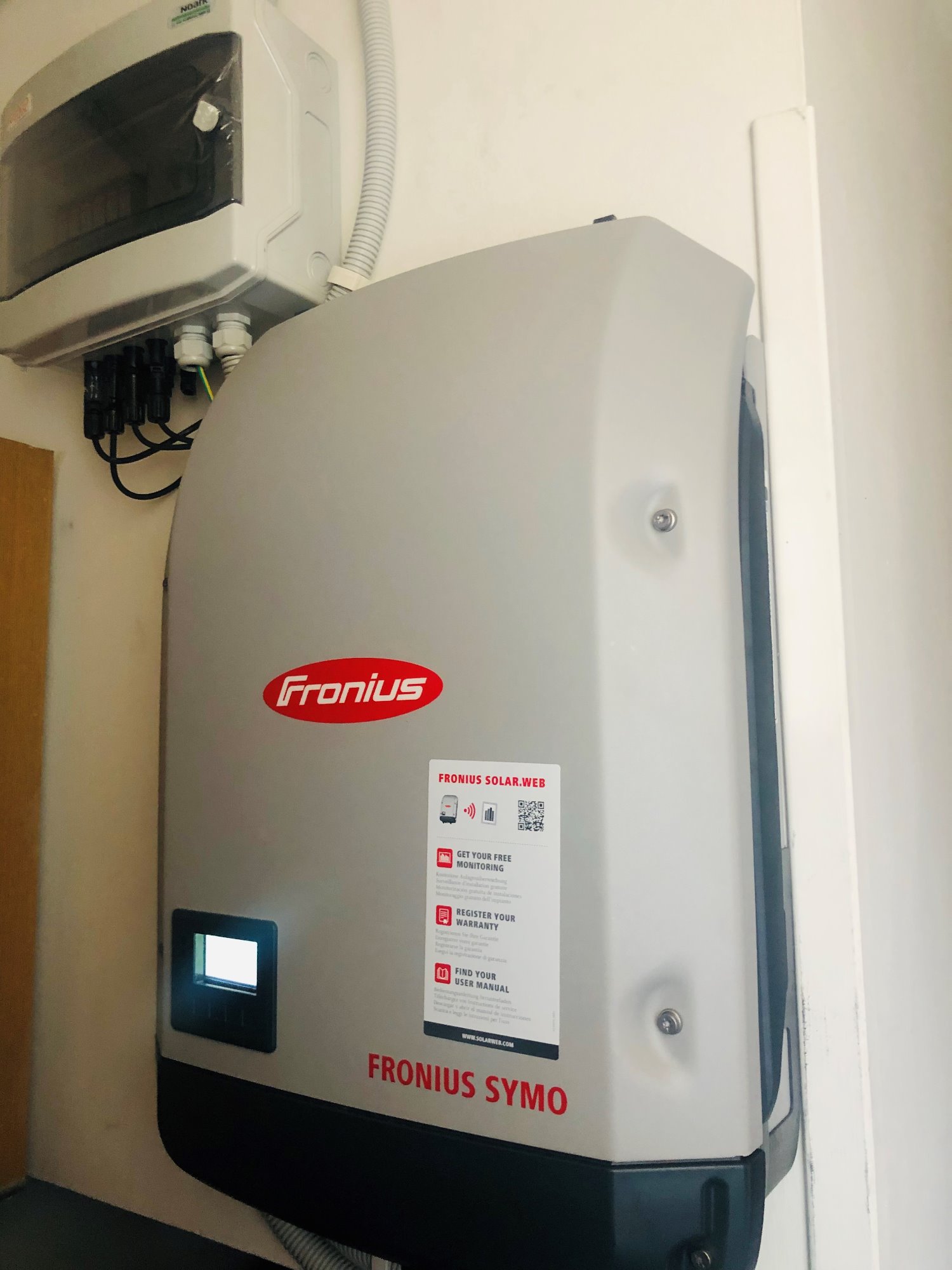 Zamontowany inwerter Fronius Symo z widocznym wyświetlaczem i naklejką informacyjną, obok puszka przyłączeniowa z przewodami.