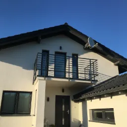 Nowoczesny dom z balkonem zabezpieczonym czarną, metalową balustradą o minimalistycznym designie, widok z dołu na tle błękitnego nieba.