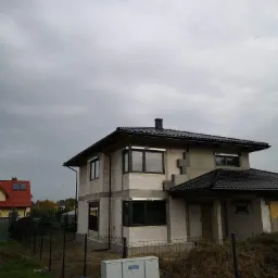 Dwukondygnacyjny dom murowany w trakcie budowy, widoczne bloczki, czarna dachówka i okna z czarną ramą, szara skrzynka elektryczna na pierwszym planie.