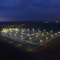 Widok z lotu ptaka na plac składowy samochodów oświetlony lampami ulicznymi, w tle widoczne zabudowania miejskie i zachodzące słońce.