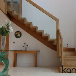 Nowoczesne schody drewniane z balustradą szklaną, zamontowane w jasnym wnętrzu z dekoracjami ściennymi i szklanym stolikiem.