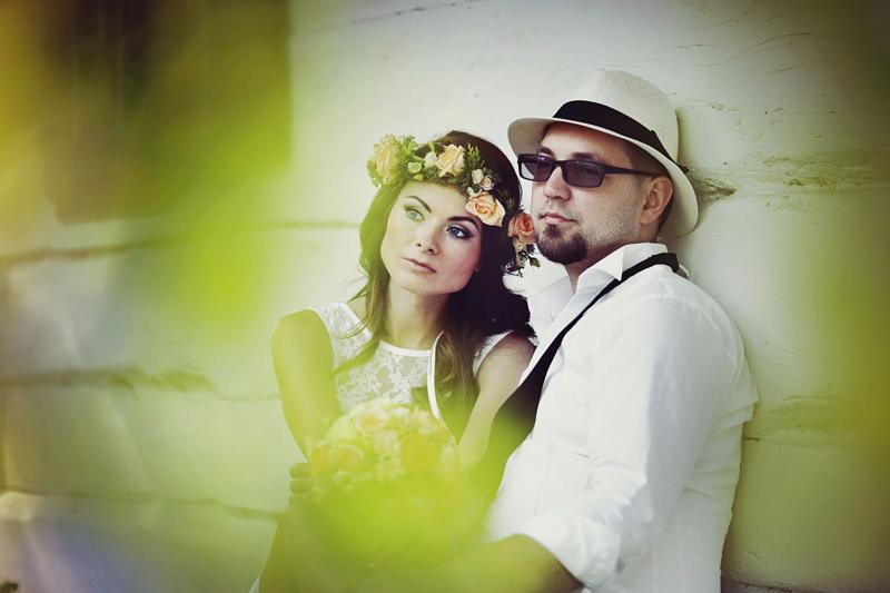 Para młoda w stylizacji rustykalnej: panna młoda w wianku z brzoskwiniowych róż, pan młody w kapeluszu i okularach przeciwsłonecznych, opierają się o białą ścianę z drewnianych bali.