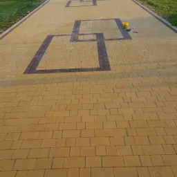 Żółta nawierzchnia z kostki brukowej ułożona we wzór geometryczny z brązowymi obramowaniami kwadratów, widoczny odpływ liniowy, paleta z kostką w tle i wiaderko na powierzchni.