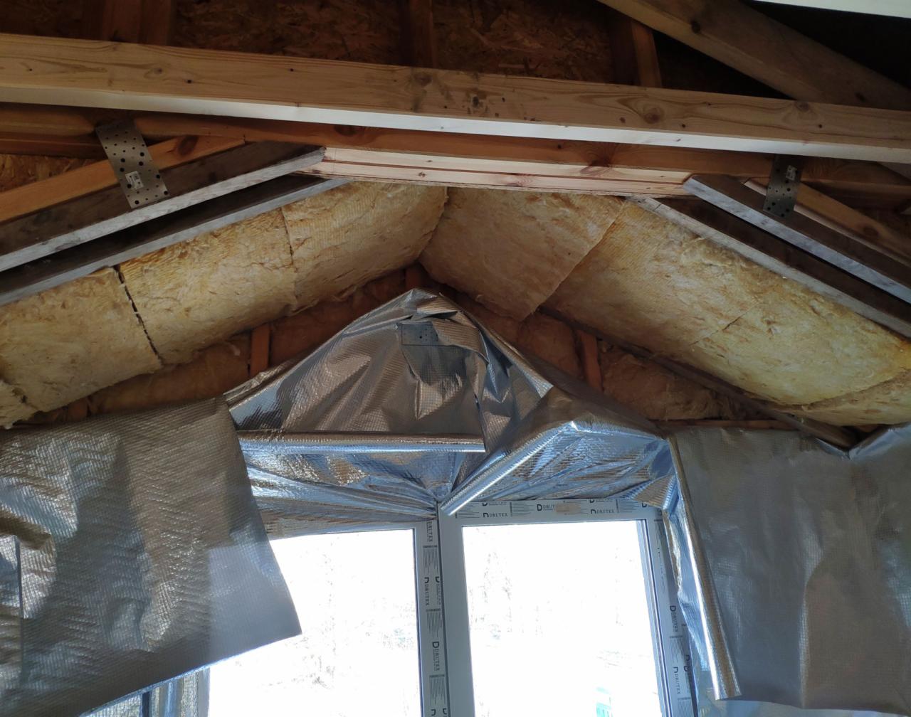 Izolacja poddasza wełną mineralną i folią aluminiową w trakcie prac wykończeniowych w domu w Warszawie, widoczne elementy konstrukcji dachu i okno.
