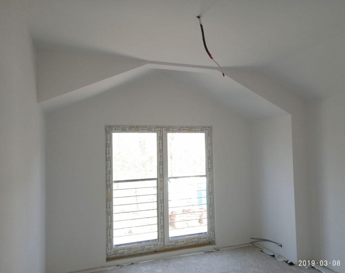 Wnętrze pokoju na poddaszu z oknem zabezpieczonym kratami, świeżo po malowaniu na biało, widoczne kable wystające z sufitu i niedokończona podłoga.
